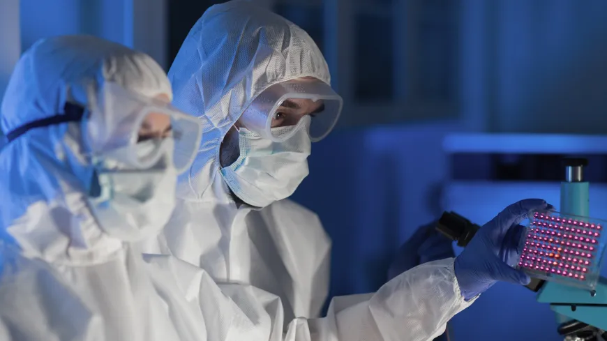 two scientists in white labcoats and white hoods with googles and masks on